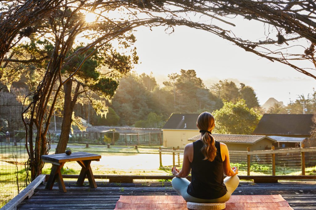 A woman sitting on a floor pillow on a deck at sunrise meditating