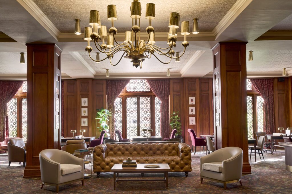 Olympic Lounge that has floor to ceiling wood paneling, a patterned ceiling, large windows with circular design in the glass, with writing desks and side chairs and leather sofa with two accents chairs and a large gold chandelier