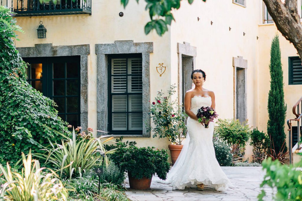 A bride in a wedding dress standing in a garden courtyard outside a Mediterranean-inspired building