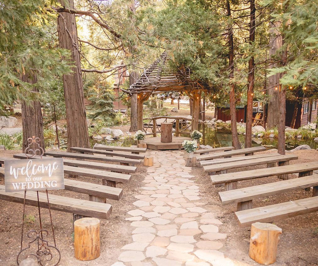 Wooden benches set for a wedding and rustic gazebo beside a koi pond beneath tall pine trees.