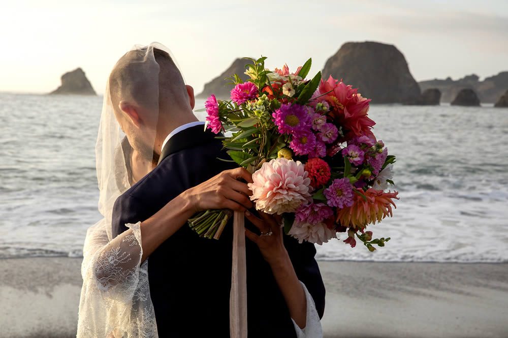 A bride and groom kissing on the beach with sea stacks in the background. The bride is holding a large wedding bouquet.