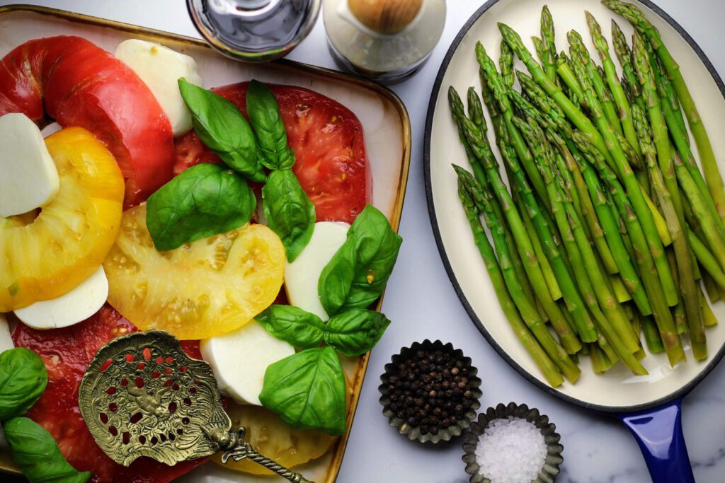 A plate of sliced tomatoes and mozzarella with basil and a plate of steam asparagus.