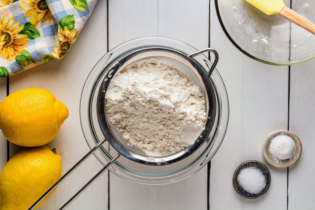 Sifting flour into a bowl with salt, baking soda and leamons set off to the side.