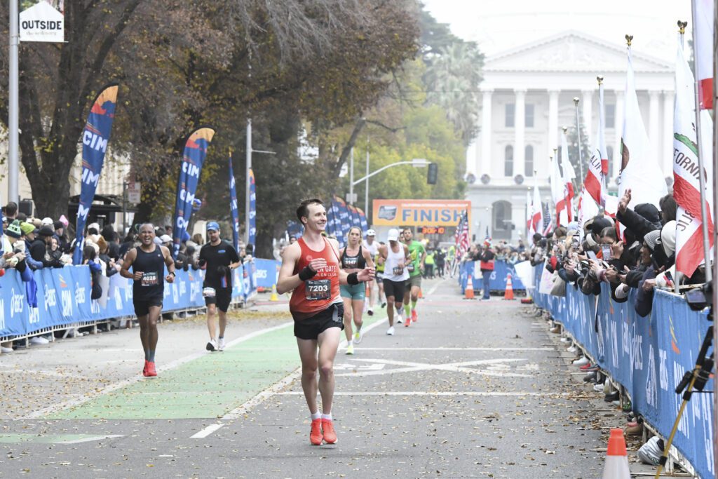 Runners passing the finish line at the California International Marathon with crowds cheering the runners on and the state capitol in the background.