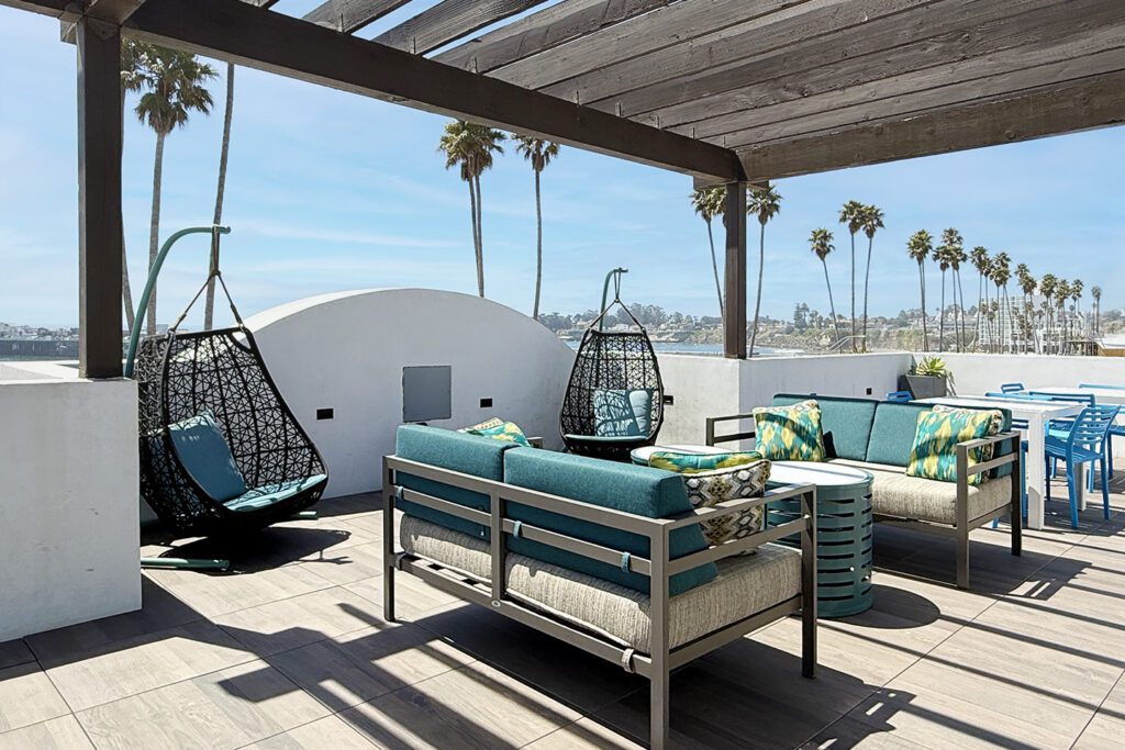 Rooftop terrace of the Marea Sol Hotel with outdoor seating, swinging chairs, and views of palm trees, the ocean, and the Santa Cruz wharf.