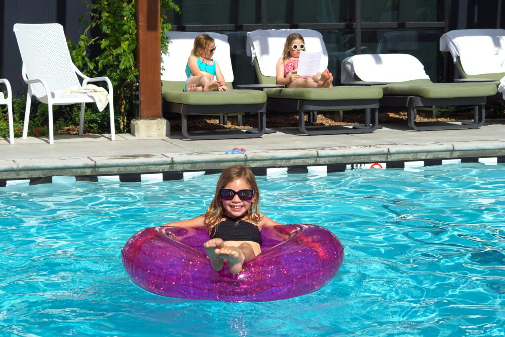 a child floating in a purple innertube in a pool with more children behind her in lounge chairs by the pool