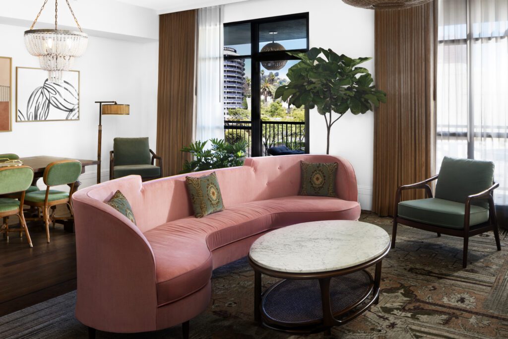 Guestroom living area with a long light pink velvet sofa, a modern armchair with light teal velvet cushion, a marble top coffee table with a dining table and velvet chairs with a beaded chandelier above and artwork hung with a floor lamp and another velvet cushioned armchair and floor to ceiling windows