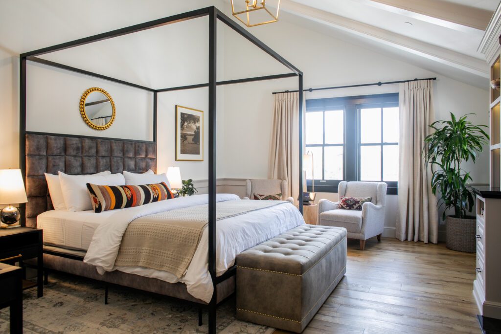Guestroom with metal canopy bed and upholstered headboard, nightstands and lamps, vaulted ceiling with a chandelier, two large windows and two accent chairs