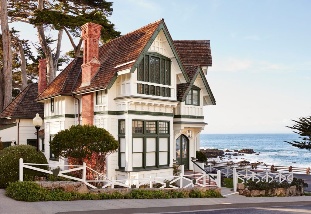 The exterior with Queen Anne Victorian architecture featuring prominent gables, decorative trim, and multi-paned windows. The inn has a white facade accented by deep forest green woodwork on the window frames and roof trim with brick chimneys extending up the side and rear of the sloping shingled roofline. A white wooden fence borders the manicured perimeter, nestled alongside Cypress trees overlooking the ocean