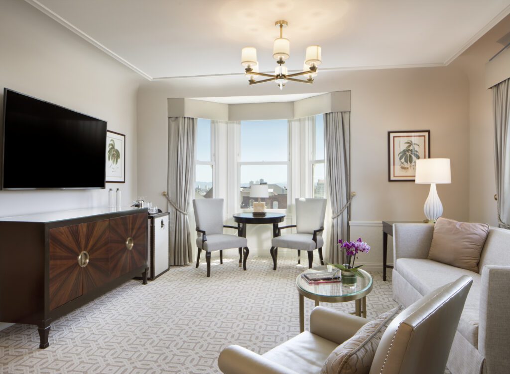 Opulent hotel suite with a table and chairs in a bay window and a couch and chair facing a flat screen television and luxurious credenza.