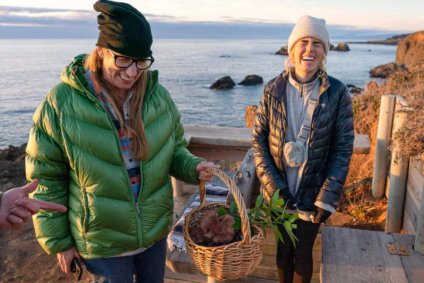Two women foraging on the coast. One is holding a basket with mushrooms and the other is laughing.